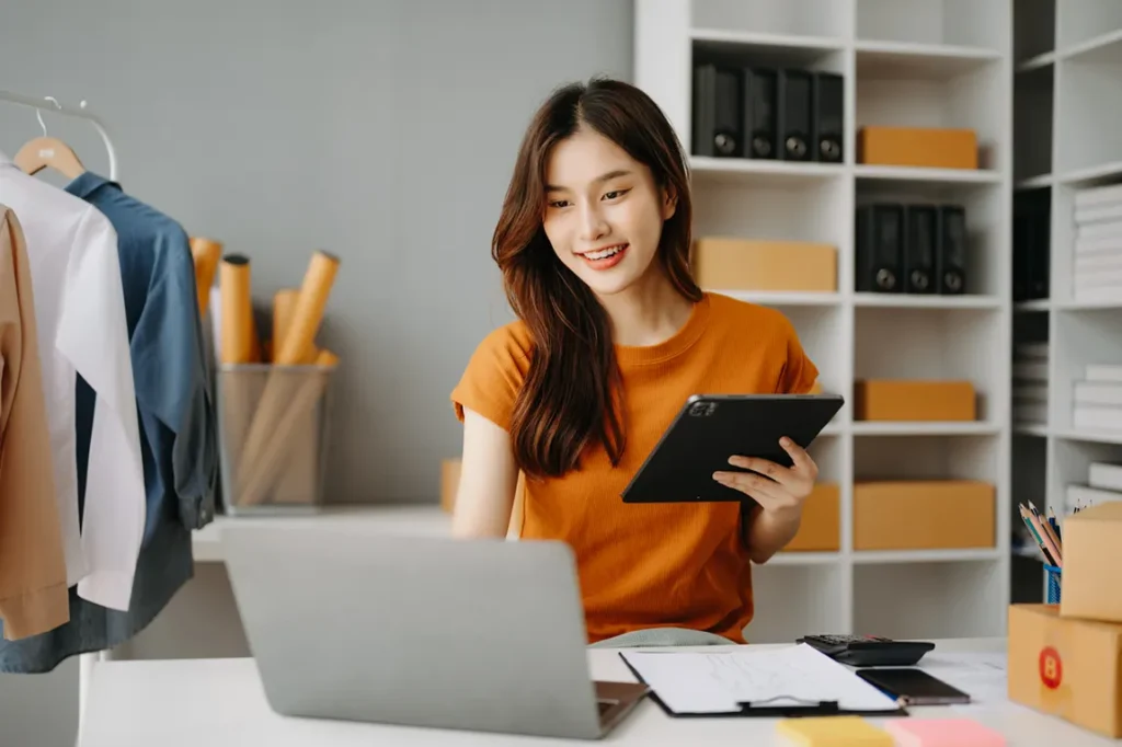 Young woman running online e-commerce business from home office with laptop, tablet, shipping boxes and clothing inventory