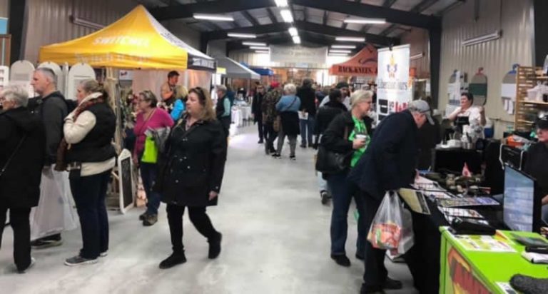 Festival marketplace scene showing vendors selling crafts and products to customers in large indoor venue space