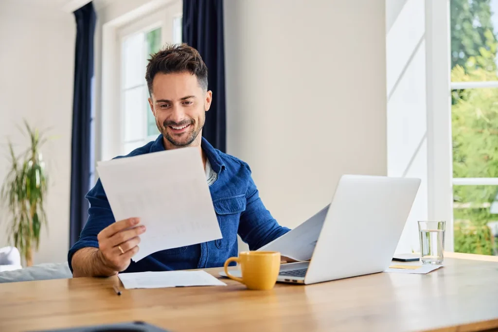 Happy businessman reviewing sole proprietorship renewal documents at home office with laptop and business paperwork