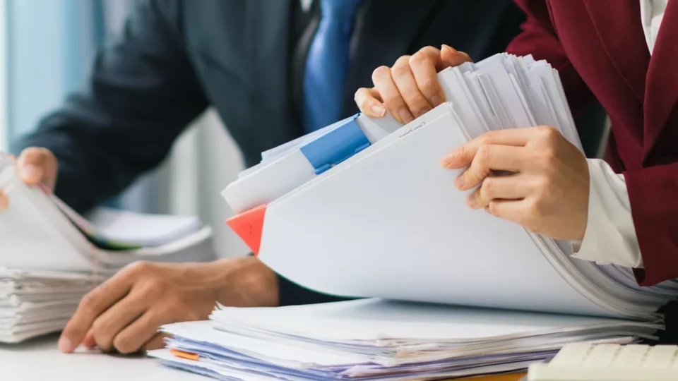 Two business professionals search through paperwork at a desk, focused on locating their corporation number.