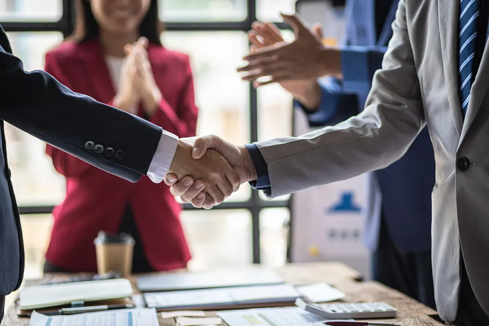 Business professionals shaking hands during partnership agreement meeting with woman applauding successful deal closure