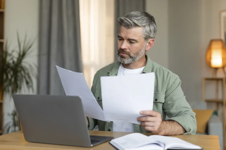Middle-aged businessman reviewing business registration documents and paperwork at home office with laptop and files