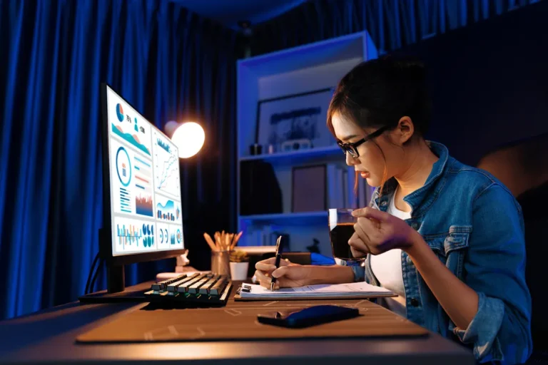 Female entrepreneur working late, reviewing analytics charts on a monitor and taking notes at a modern home office desk.