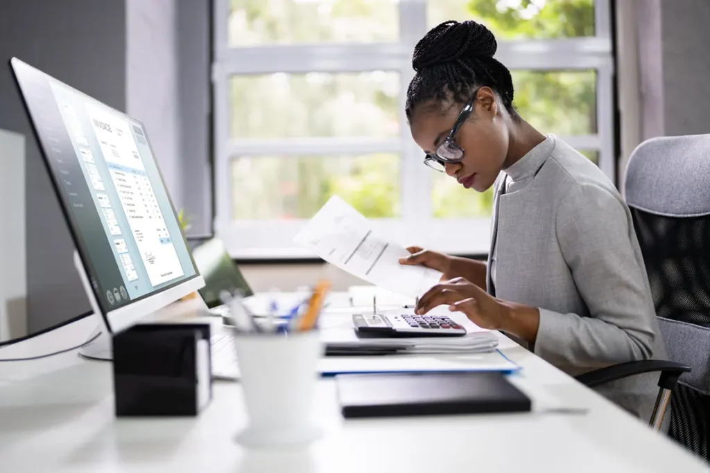 Professional woman working on invoices at desk with calculator and computer, managing side hustle payments and bookkeeping