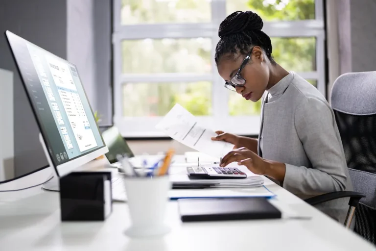 Professional woman working on invoices at desk with calculator and computer, managing side hustle payments and bookkeeping