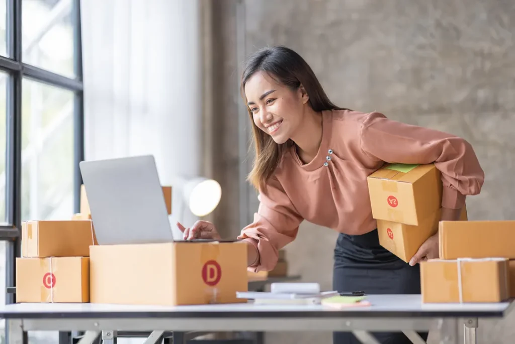 Smiling entrepreneur managing her side hustle packaging orders while checking her laptop in a small home office