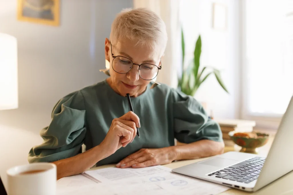 An older woman with glasses sits at a desk, using a laptop to search for information about a business.