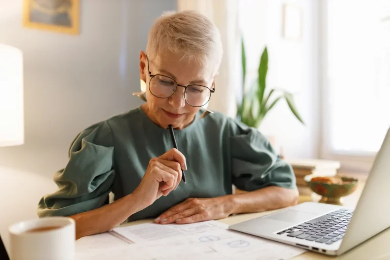 An older woman with glasses sits at a desk, using a laptop to search for information about a business.