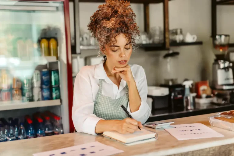 A woman wearing an apron standing behind a counter reviews registration options in a store or café setting.