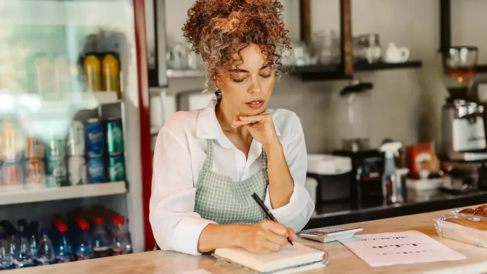 A woman wearing an apron standing behind a counter reviews registration options in a store or café setting.